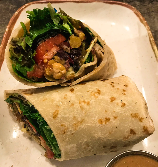 The Bella sandwich from Mudgie's Deli in Detroit, MI features tomato, chick peas, roasted red pepper, artichoke hearts, olive tapenade, sunflower sprouts, mixed greens, sunflower seeds, rolled up in a flatbread with a balsamic vinaigrette. 